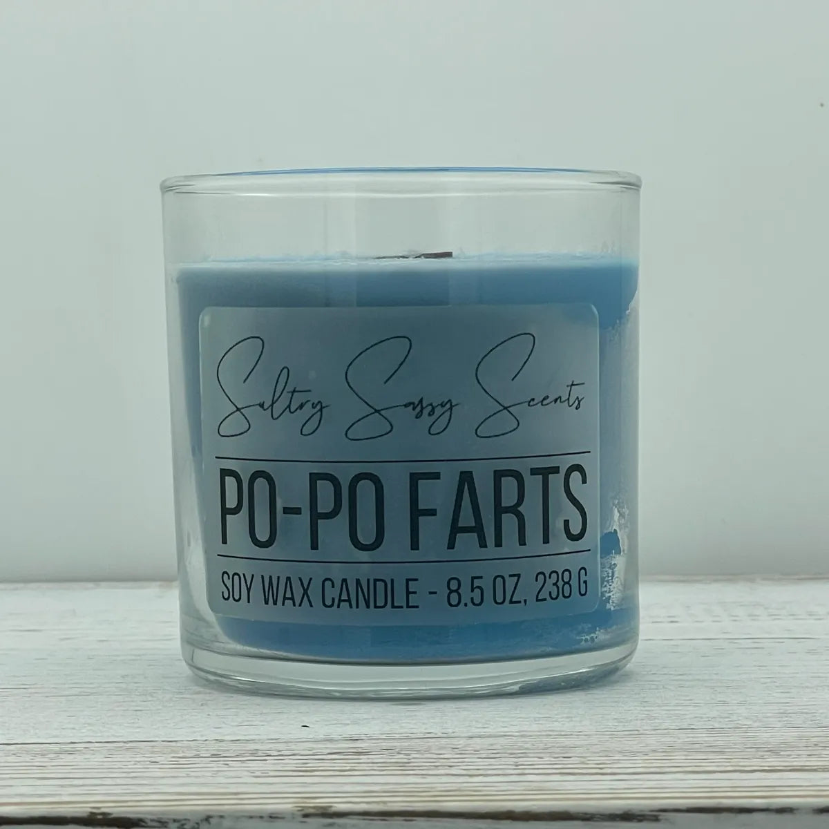 An 8.5-ounce clear glass jar of Sultry Sassy Scents "Po-Po Farts" soy wax candle displaying a dusty blue wax color and a wooden wick, centered on a light wood surface.