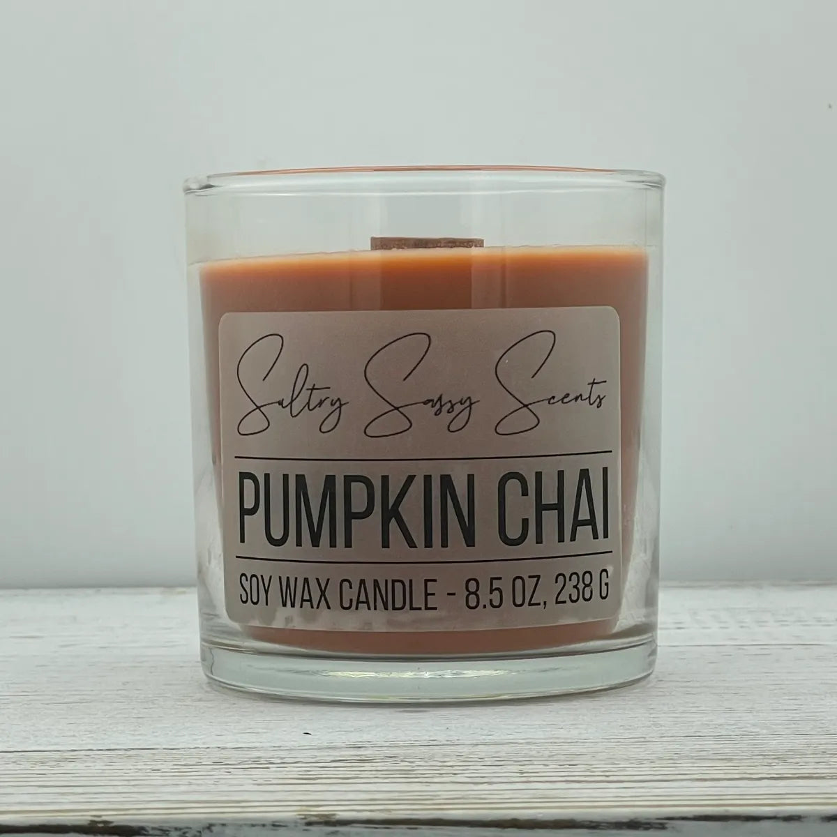 An 8.5-ounce clear glass jar of Sultry Sassy Scents "Pumpkin Chai" soy wax candle showcasing a warm terracotta orange wax color with a wooden wick, resting on a white-washed wood table.