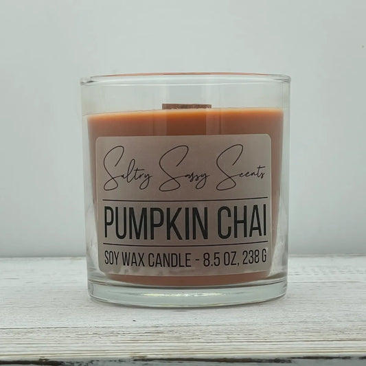 An 8.5-ounce clear glass jar of Sultry Sassy Scents "Pumpkin Chai" soy wax candle showcasing a warm terracotta orange wax color with a wooden wick, resting on a white-washed wood table.