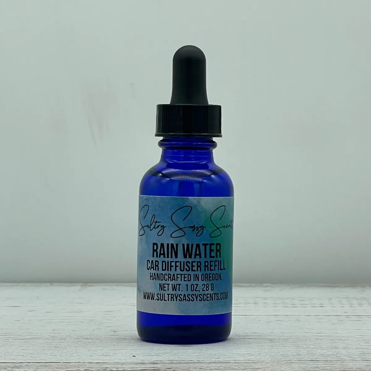 Front label view of a cobalt blue glass dropper bottle containing Sultry Sassy Scents "Rain Water" Car Diffuser Refill, detailing the 1 ounce net weight.