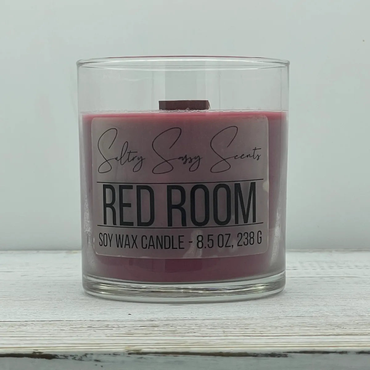 An 8.5-ounce clear glass jar of Sultry Sassy Scents "Red Room" soy wax candle displaying a muted plum red wax color and a wooden wick, showcased on a light wooden background.