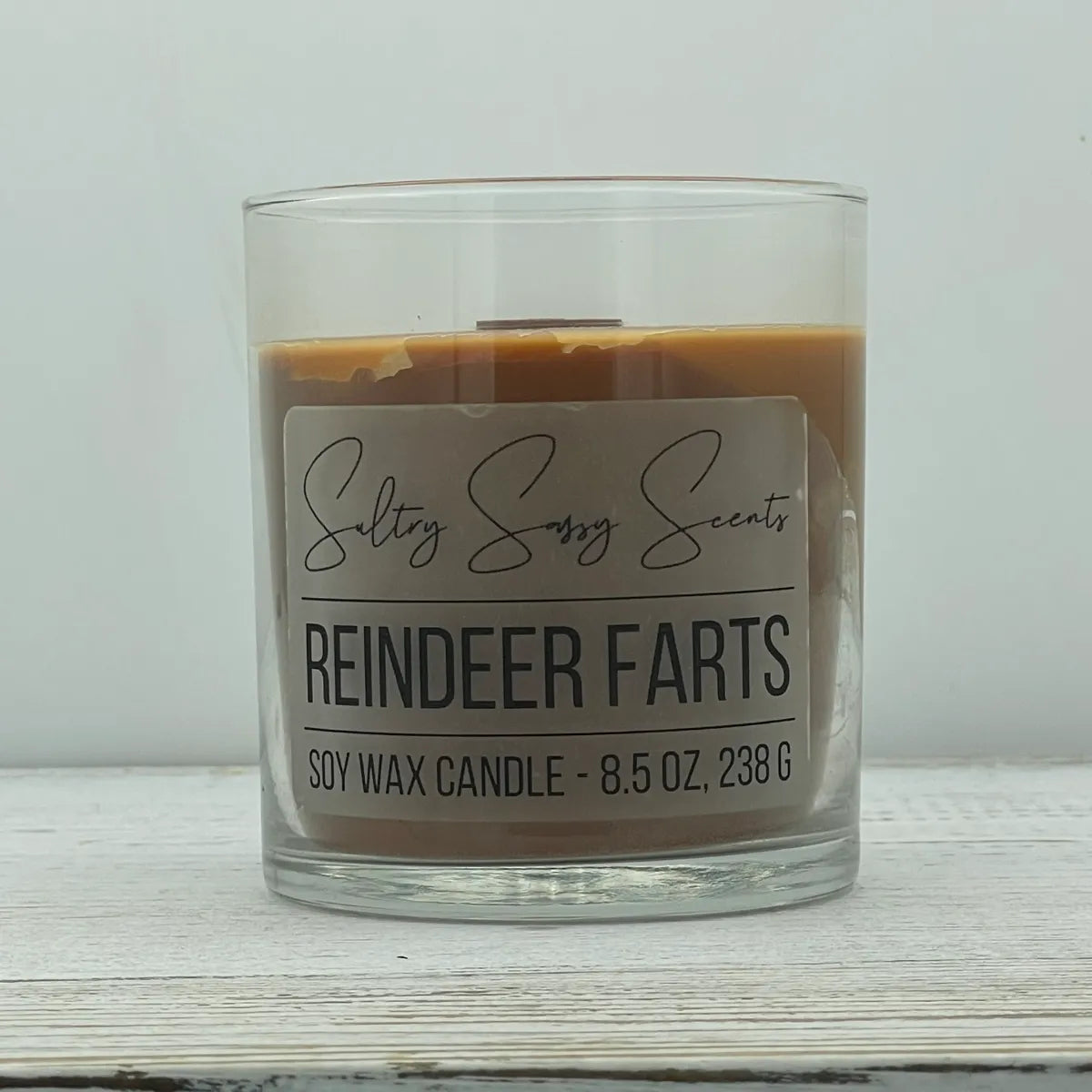 An 8.5-ounce clear glass jar of Sultry Sassy Scents "Reindeer Farts" soy wax candle featuring a smooth milk chocolate brown wax color, resting on a white wood surface.
