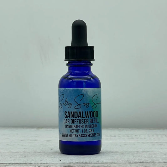 Front label view of a cobalt blue glass dropper bottle containing Sultry Sassy Scents "Sandalwood" Car Diffuser Refill, centered against a light gray backdrop.