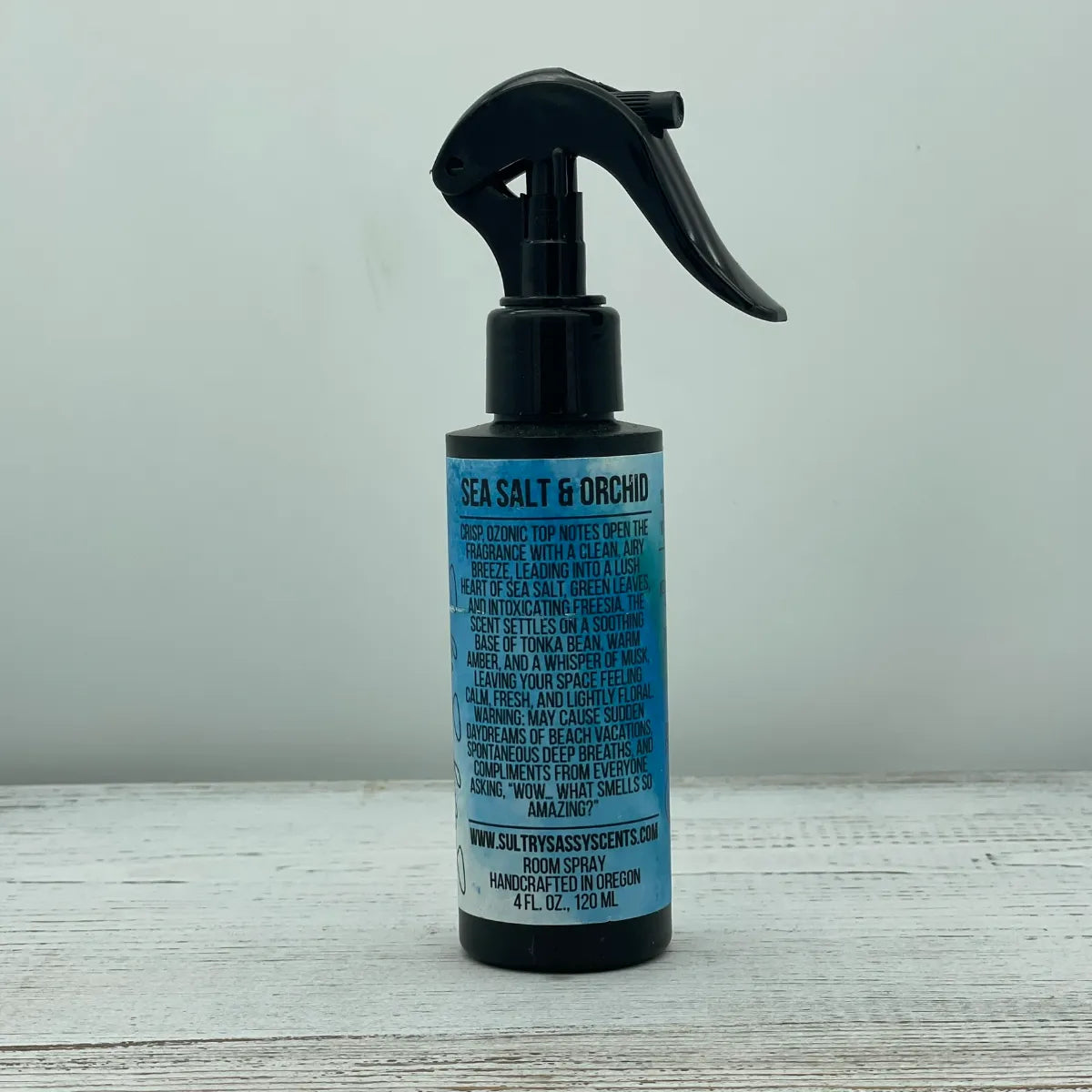 Front label of a black spray bottle for Sultry Sassy Scents "Sea Salt & Orchid" Room Spray, showcasing a paragraph block description of a crisp, airy breeze fragrance with tonka bean, amber, and musk notes.