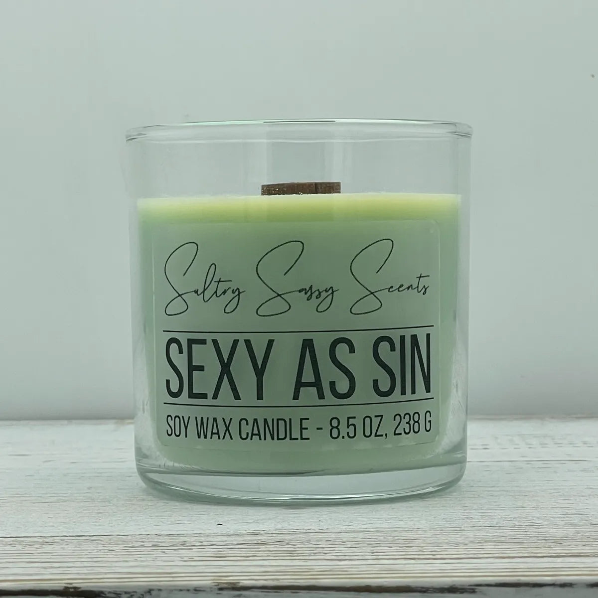 An 8.5-ounce clear glass jar of Sultry Sassy Scents "Sexy As Sin" soy wax candle displaying a light pastel sage green wax color and a wooden wick, centered on a white wood background.