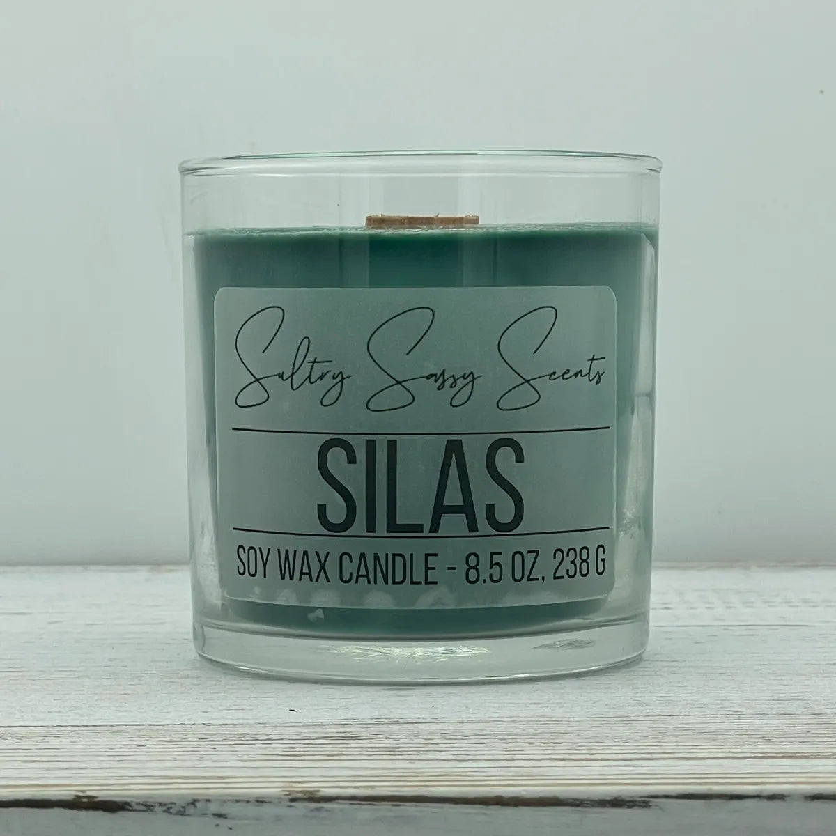 An 8.5-ounce clear glass jar of Sultry Sassy Scents "Silas" soy wax candle showcasing a deep forest green wax color with a flat wooden wick, displayed on a rustic light wood table.