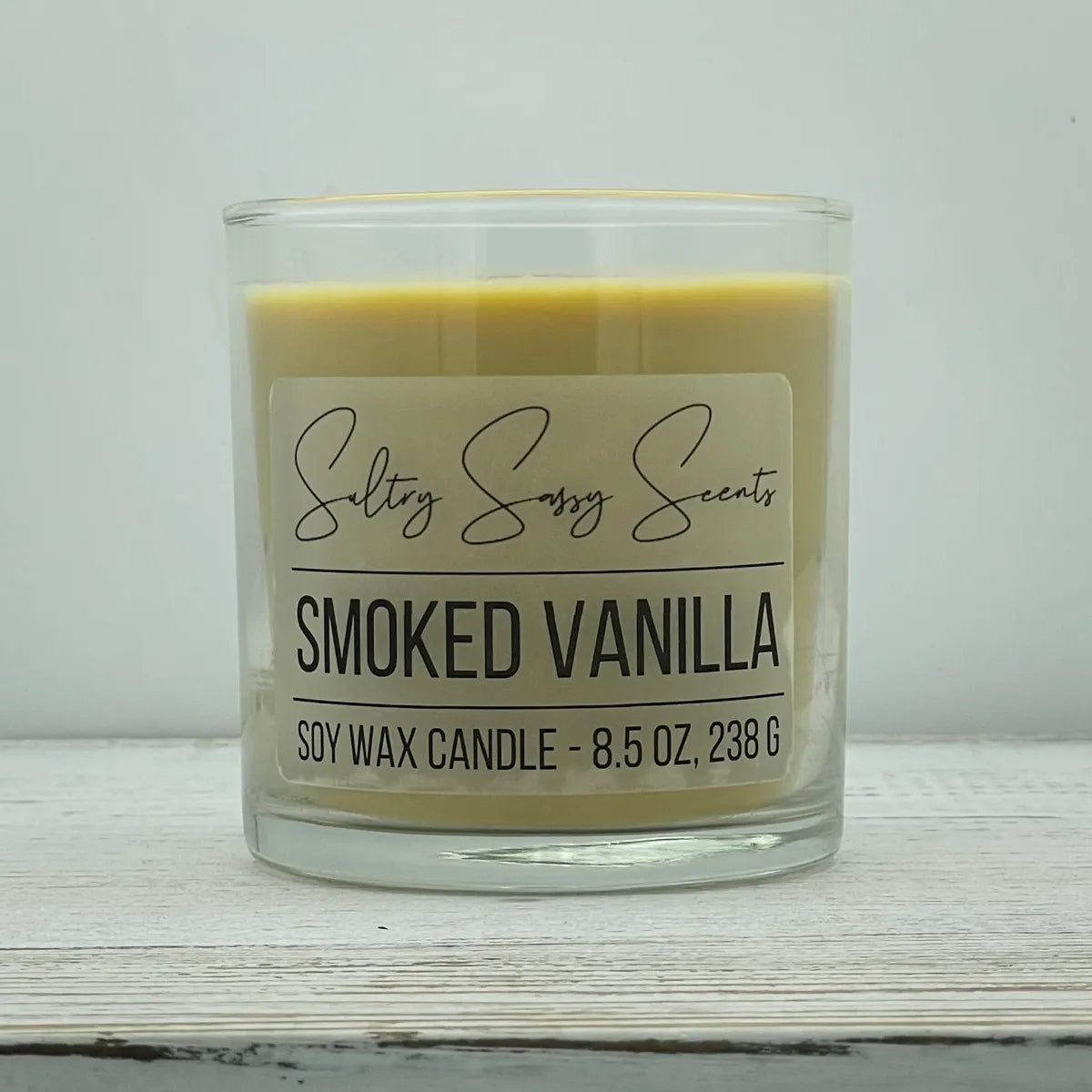An 8.5-ounce clear glass jar of Sultry Sassy Scents "Smoked Vanilla" soy wax candle featuring a smooth cream-colored wax, centered on a rustic white wood background.