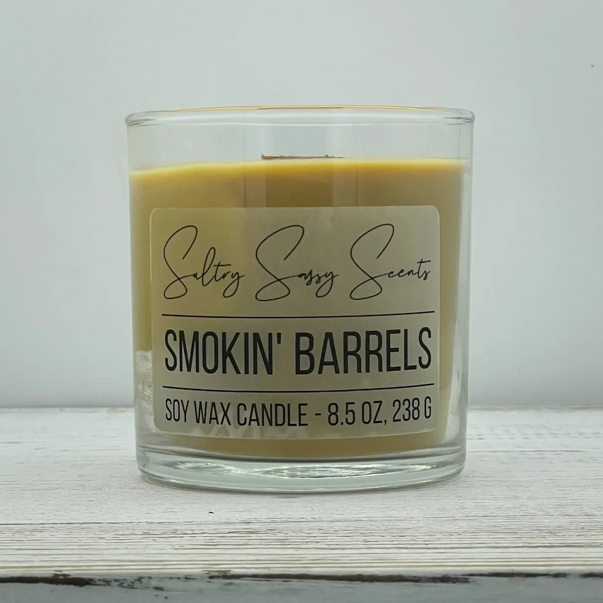An 8.5-ounce clear glass jar of Sultry Sassy Scents "Smokin' Barrels" soy wax candle showcasing a golden amber wax color and a wooden wick, displayed on a light wooden background.