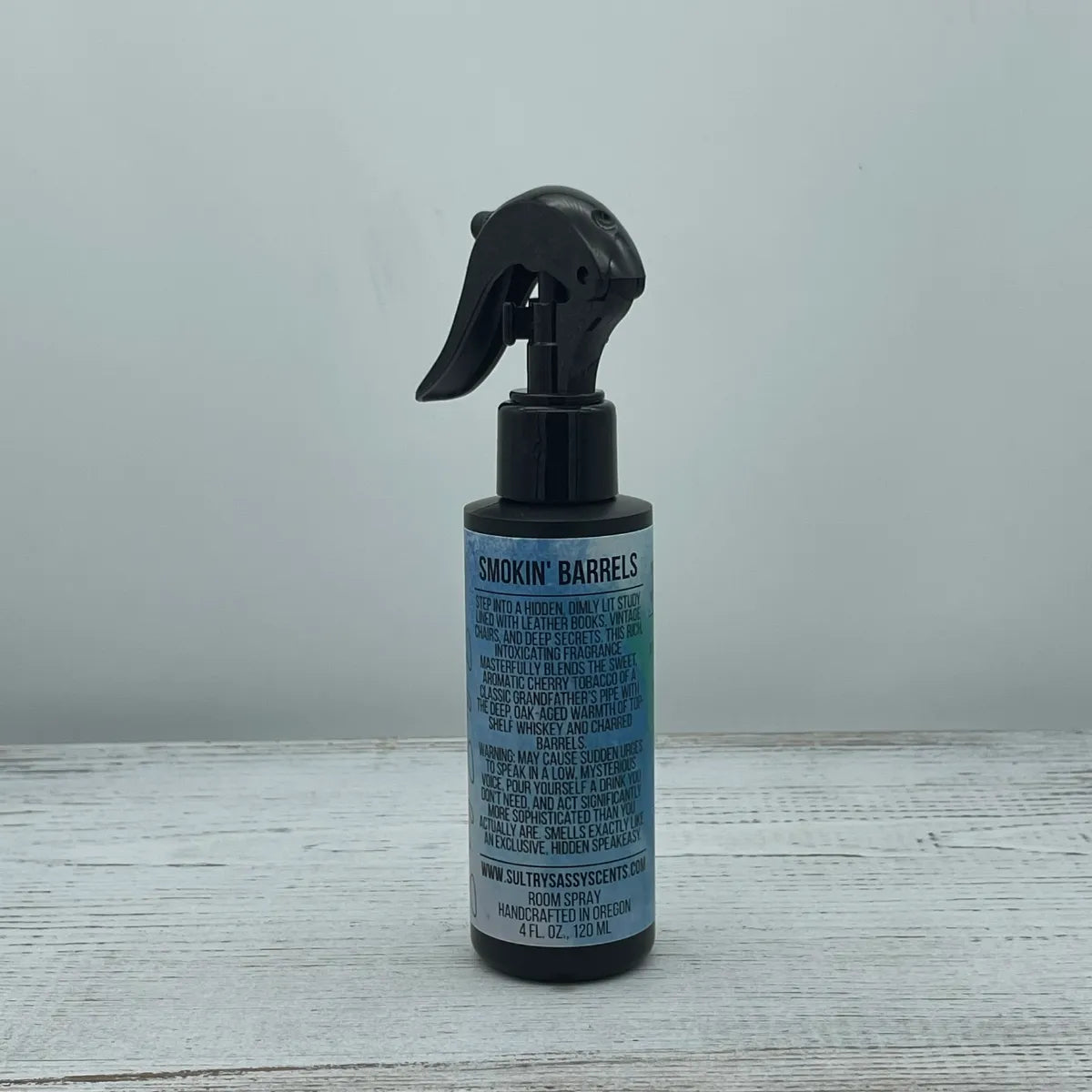 Front label view of a 4-ounce black spray bottle for Sultry Sassy Scents "Smokin' Barrels" Room Spray, showing a blue-toned label detailing handcrafted in Oregon information on a white wooden table.