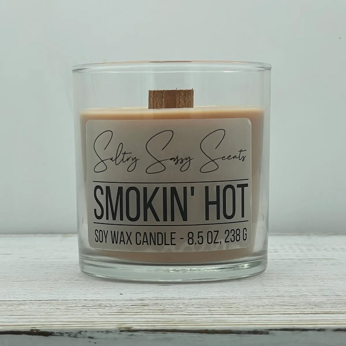 An 8.5-ounce clear glass jar of Sultry Sassy Scents "Smokin' Hot" soy wax candle featuring a soft warm taupe wax color with a flat wooden wick, positioned against a clean light background.