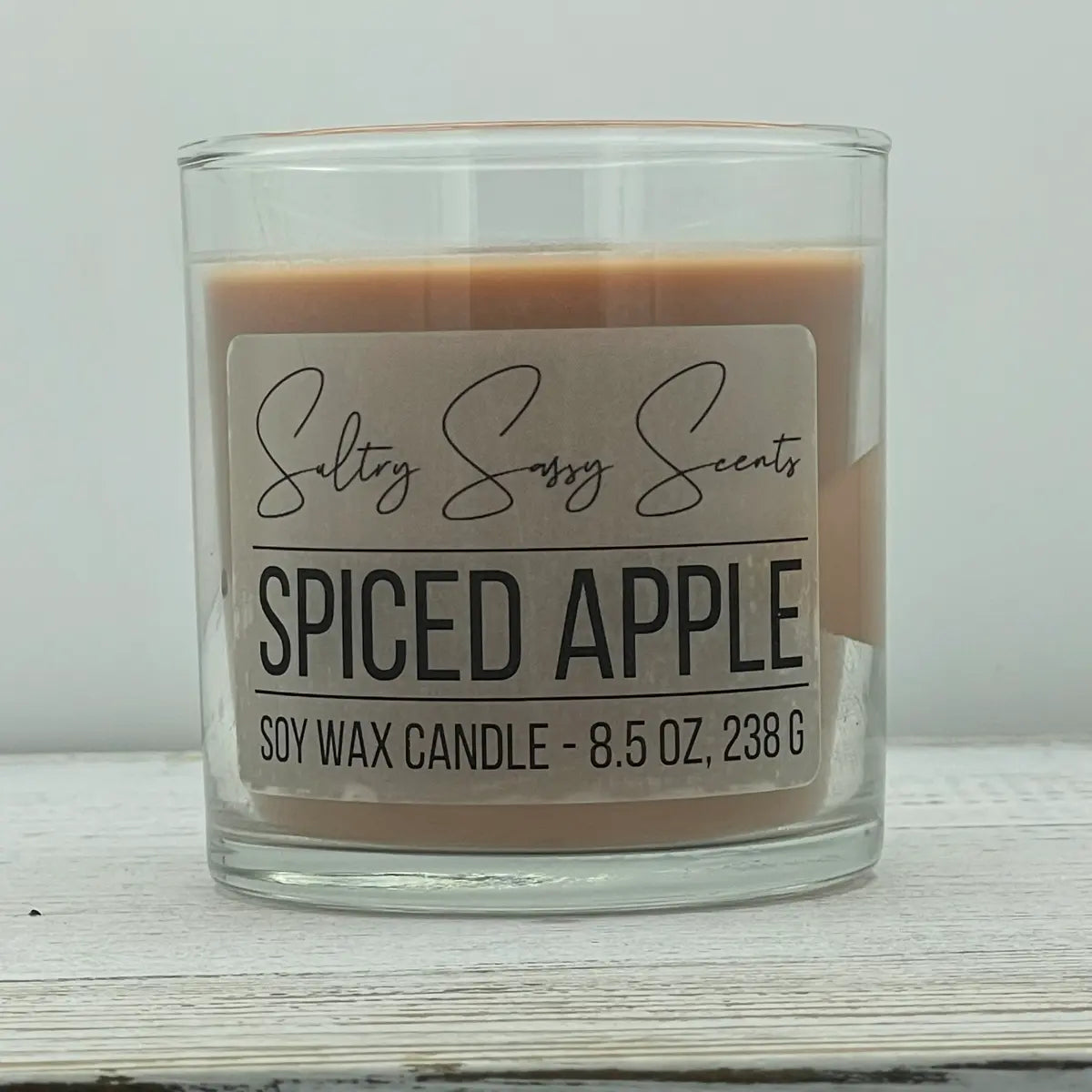 An 8.5-ounce clear glass jar of Sultry Sassy Scents "Spiced Apple" soy wax candle showcasing a warm, muted tan-brown wax color and a wooden wick.