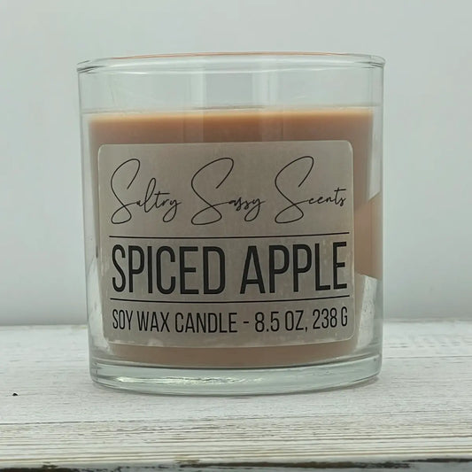 An 8.5-ounce clear glass jar of Sultry Sassy Scents "Spiced Apple" soy wax candle showcasing a warm, muted tan-brown wax color and a wooden wick.