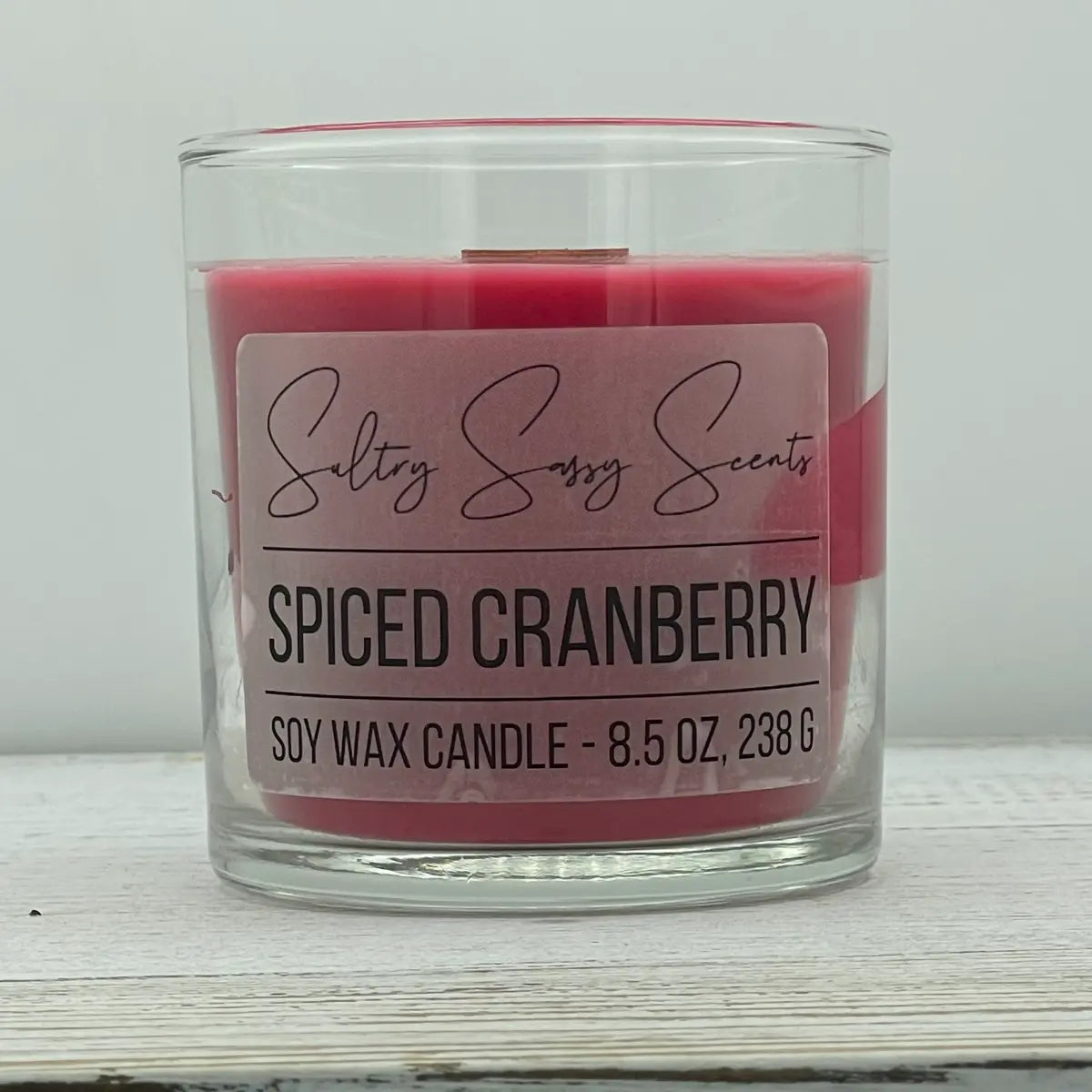 An 8.5-ounce clear glass jar of Sultry Sassy Scents "Spiced Cranberry" soy wax candle featuring a rich, solid dark pink wax color and a flat wooden wick.