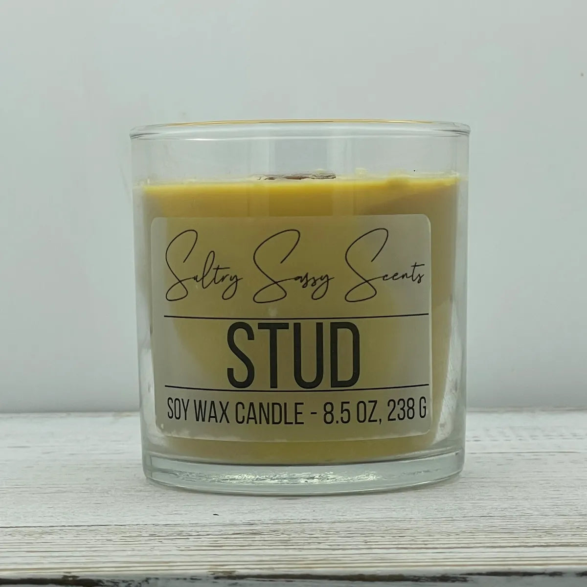 An 8.5-ounce clear glass jar of Sultry Sassy Scents "Stud" soy wax candle displaying a golden tan wax color and a wooden wick, resting on a white-washed wood table.
