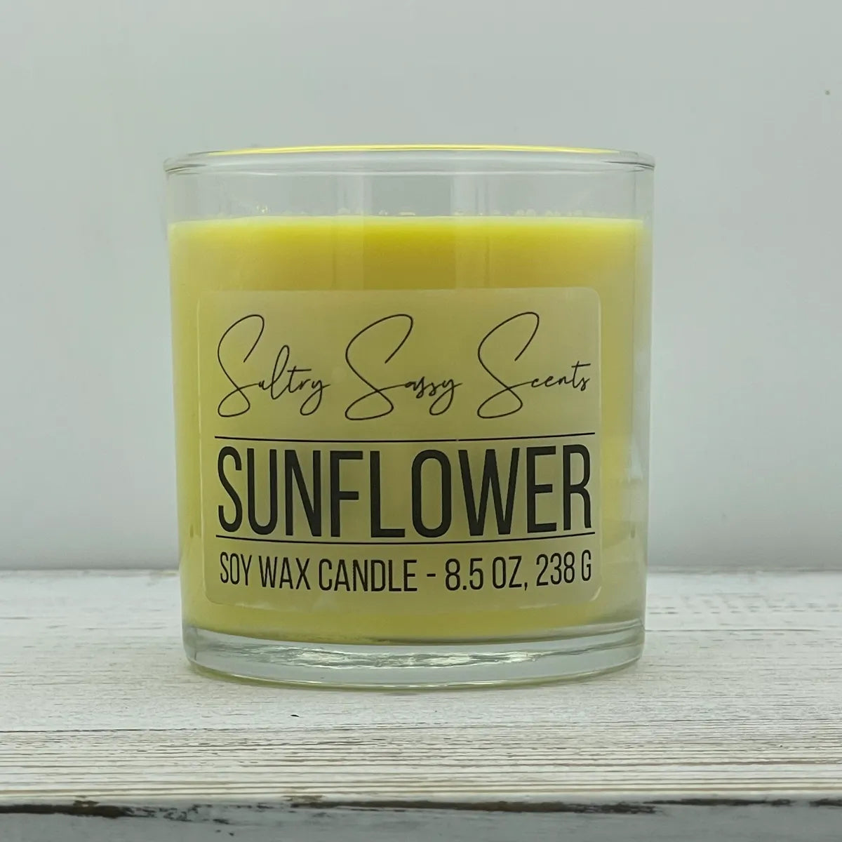 An 8.5-ounce clear glass jar of Sultry Sassy Scents "Sunflower" soy wax candle showcasing a pastel yellow wax color, resting on a white wooden surface.