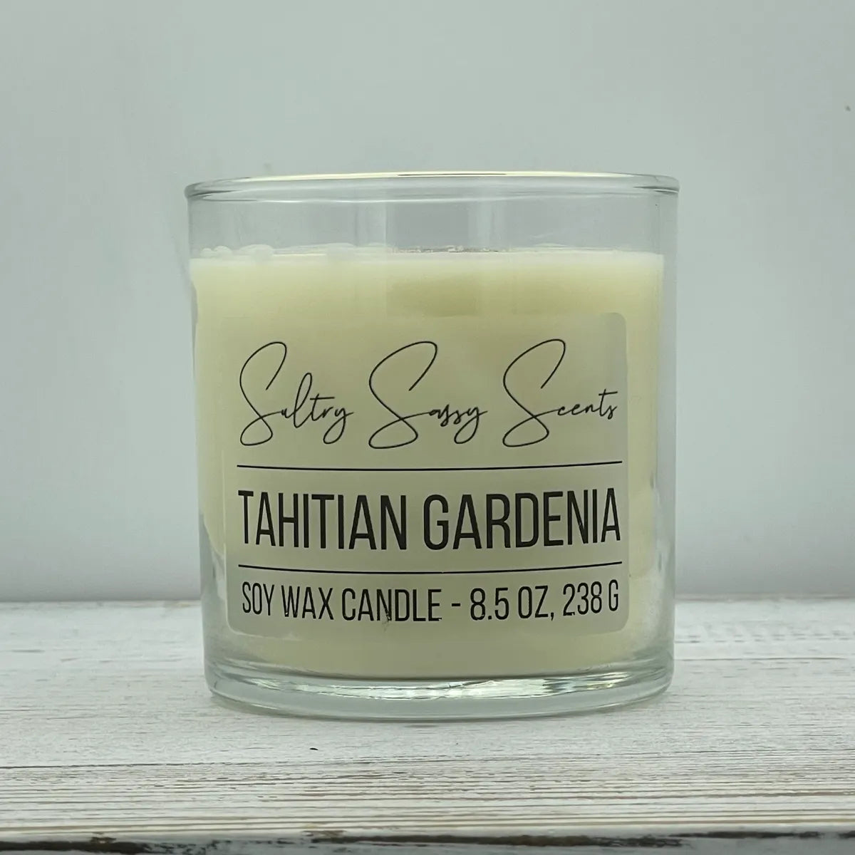An 8.5-ounce clear glass jar of Sultry Sassy Scents "Tahitian Gardenia" soy wax candle featuring a classic off-white wax color, displayed on a light wooden background.