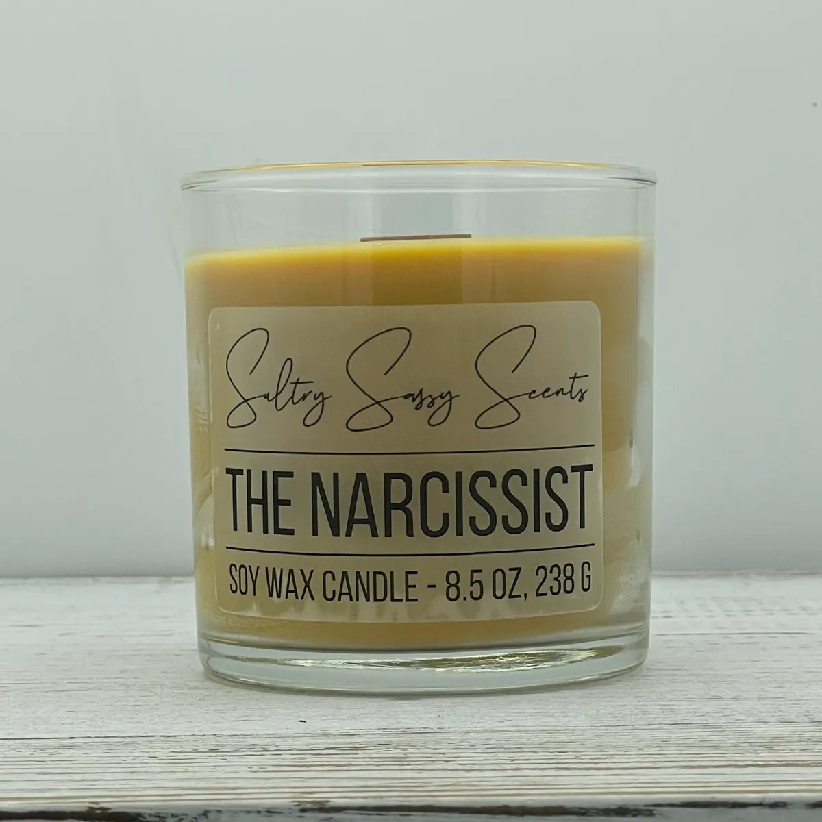 An 8.5-ounce clear glass jar of Sultry Sassy Scents "The Narcissist" soy wax candle featuring a warm mustard yellow wax color and a flat wooden wick, centered on a white wood backdrop.