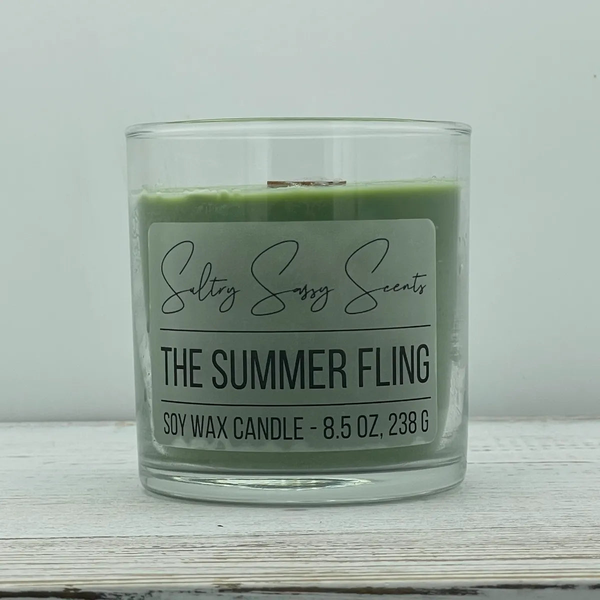 An 8.5-ounce clear glass jar of Sultry Sassy Scents "The Summer Fling" soy wax candle featuring a muted olive green wax color and a wooden wick, displayed against a clean light background.