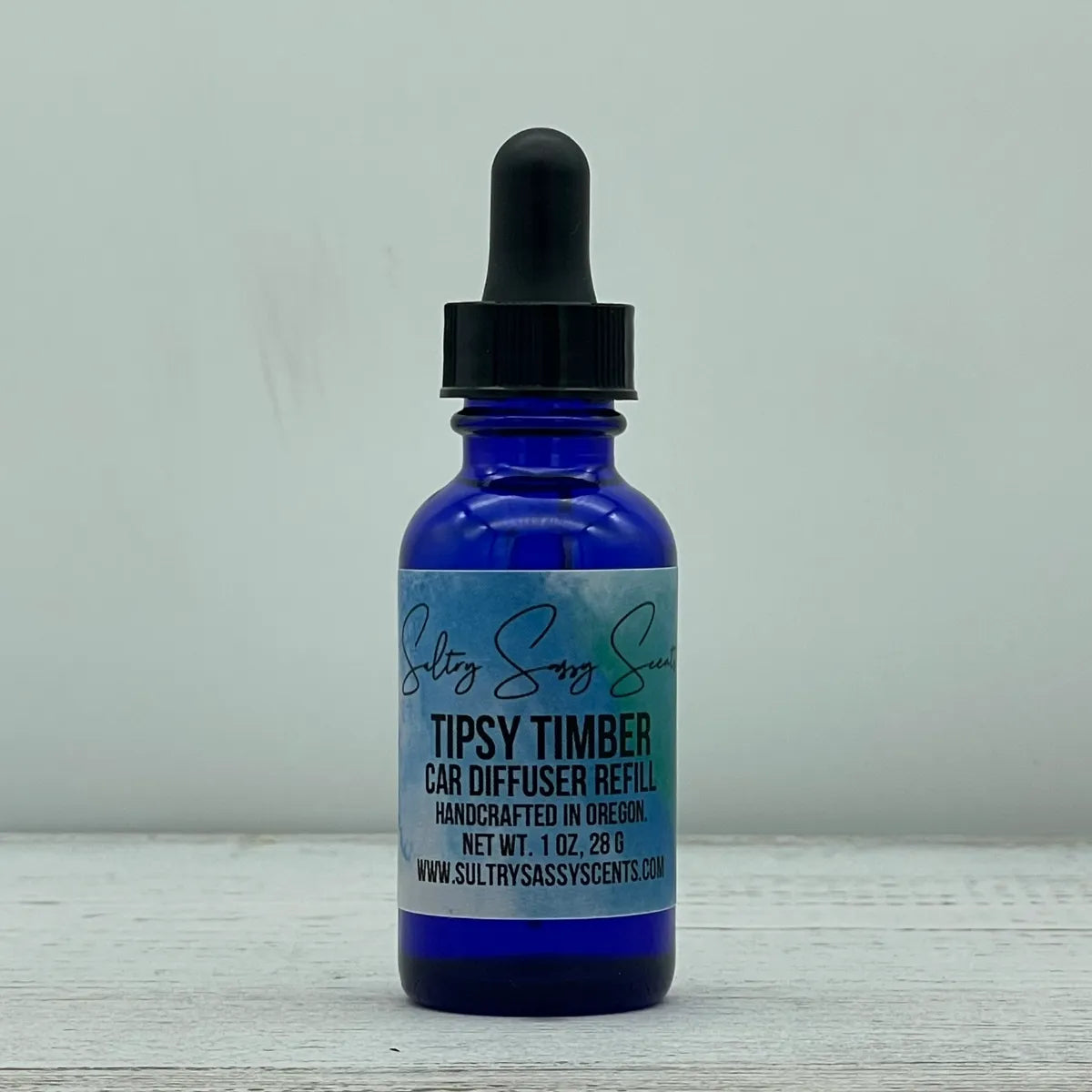 Front label view of a cobalt blue glass dropper bottle containing Sultry Sassy Scents "Tipsy Timber" Car Diffuser Refill, resting on a rustic white wood table.
