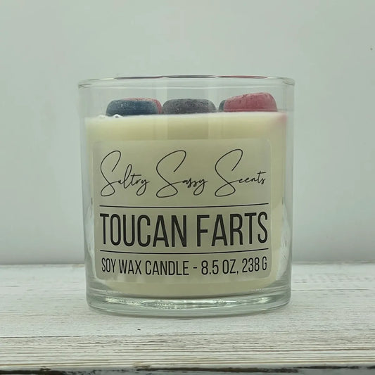 An 8.5-ounce clear glass jar of Sultry Sassy Scents "Toucan Farts" soy wax candle featuring off-white wax decorated with three embedded colorful fruit loop wax toppers.
