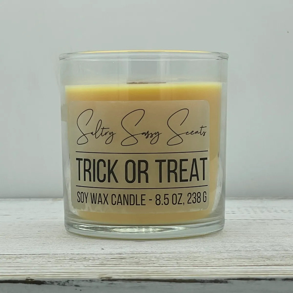 An 8.5-ounce clear glass jar of Sultry Sassy Scents "Trick or Treat" soy wax candle showcasing a smooth pastel orange wax color and a wooden wick, centered against a light gray background.