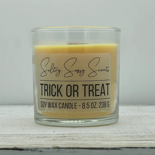 An 8.5-ounce clear glass jar of Sultry Sassy Scents "Trick or Treat" soy wax candle showcasing a smooth pastel orange wax color and a wooden wick, centered against a light gray background.