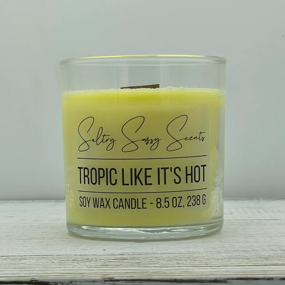 An 8.5-ounce clear glass jar of Sultry Sassy Scents "Tropic Like It's Hot" soy wax candle highlighting a soft pastel yellow wax color and a wooden wick, displayed on a rustic wood surface.