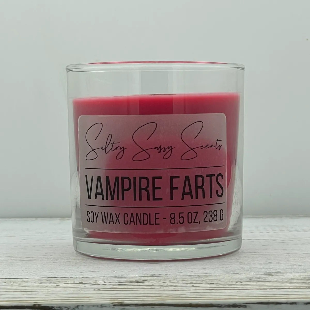 An 8.5-ounce clear glass jar of Sultry Sassy Scents "Vampire Farts" soy wax candle featuring a deep crimson red wax color, displayed on a light wooden background.