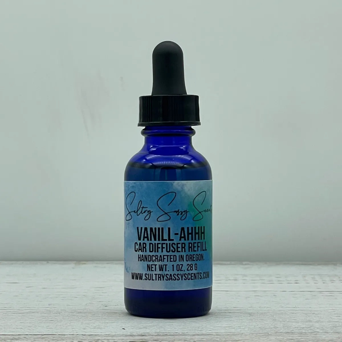 Front label view of a cobalt blue glass dropper bottle containing Sultry Sassy Scents "Vanill-Ahhh" Car Diffuser Refill, highlighting the bold product name layout on a white wood surface.