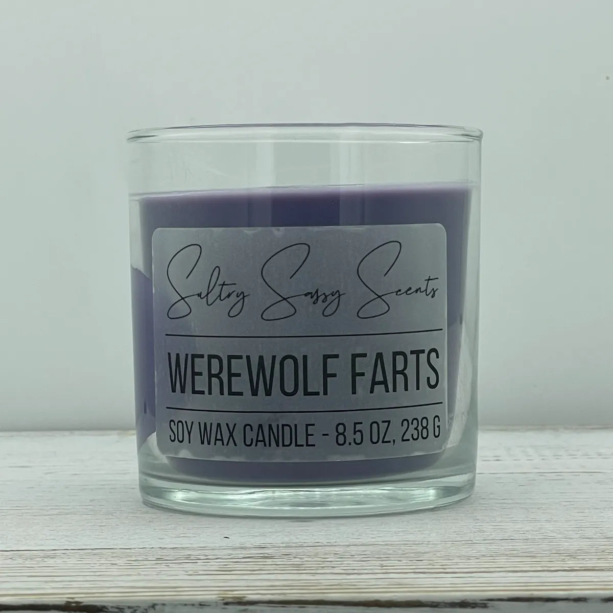 An 8.5-ounce clear glass jar of Sultry Sassy Scents "Werewolf Farts" soy wax candle featuring a deep, dark purple wax color, displayed on a rustic white wood background.