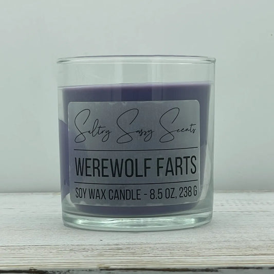 An 8.5-ounce clear glass jar of Sultry Sassy Scents "Werewolf Farts" soy wax candle featuring a deep, dark purple wax color, displayed on a rustic white wood background.