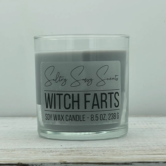 An 8.5-ounce clear glass jar of Sultry Sassy Scents "Witch Farts" soy wax candle displaying a muted charcoal gray wax color and a wooden wick.