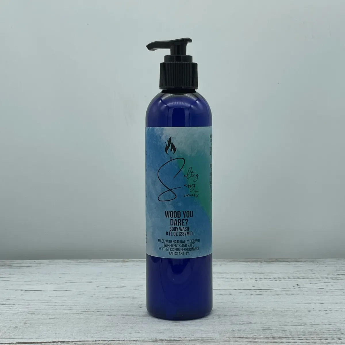 Front label view of a blue pump bottle containing Sultry Sassy Scents "Wood You Dare?" Body Wash, showcasing the product name and branding text over a stylized watercolor background.