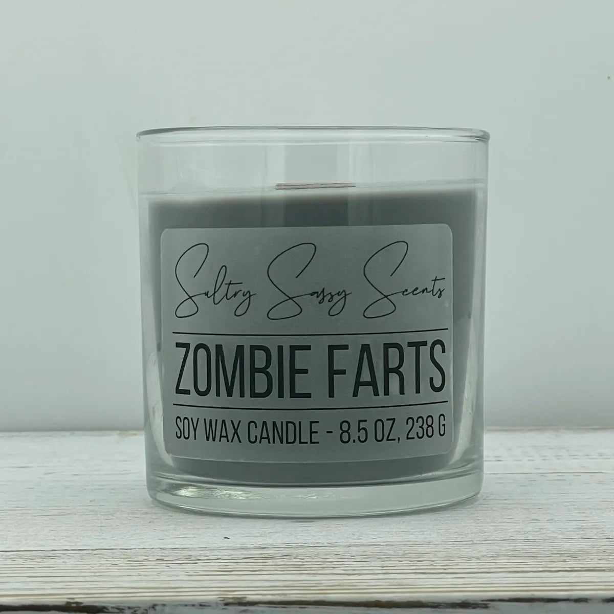 An 8.5-ounce clear glass jar of Sultry Sassy Scents "Zombie Farts" soy wax candle showcasing a smooth dark charcoal gray wax color with a flat wooden wick.