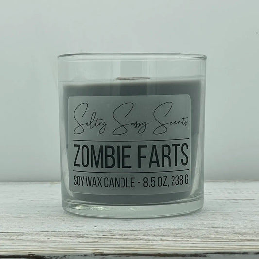 An 8.5-ounce clear glass jar of Sultry Sassy Scents "Zombie Farts" soy wax candle showcasing a smooth dark charcoal gray wax color with a flat wooden wick.